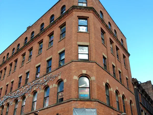 Red brick Victorian corner building with rows of arched windows, poster-covered ground-floor shopfronts and an arched doorway, under a clear blue sky. — Afflecks, Manchester