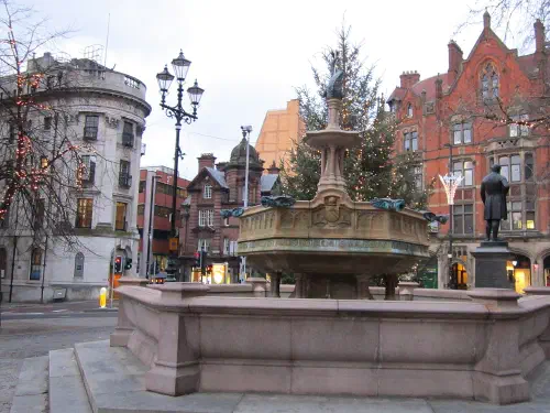 Fountain in town square with Christmas tree, historic buildings, bare branches, and overcast sky.
