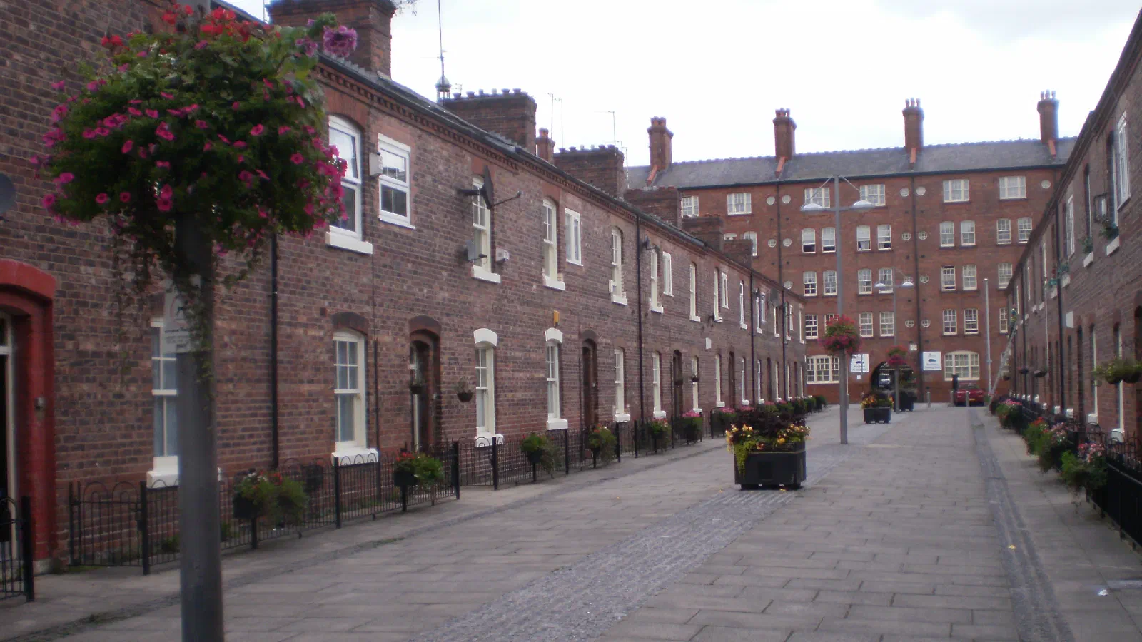 Red brick houses line a quiet stone street decorated with hanging flowers under an overcast sky. — Anita Street, Manchester