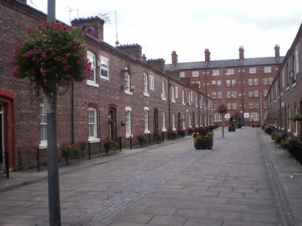 Red brick houses line a quiet stone street decorated with hanging flowers under an overcast sky. — Anita Street, Manchester