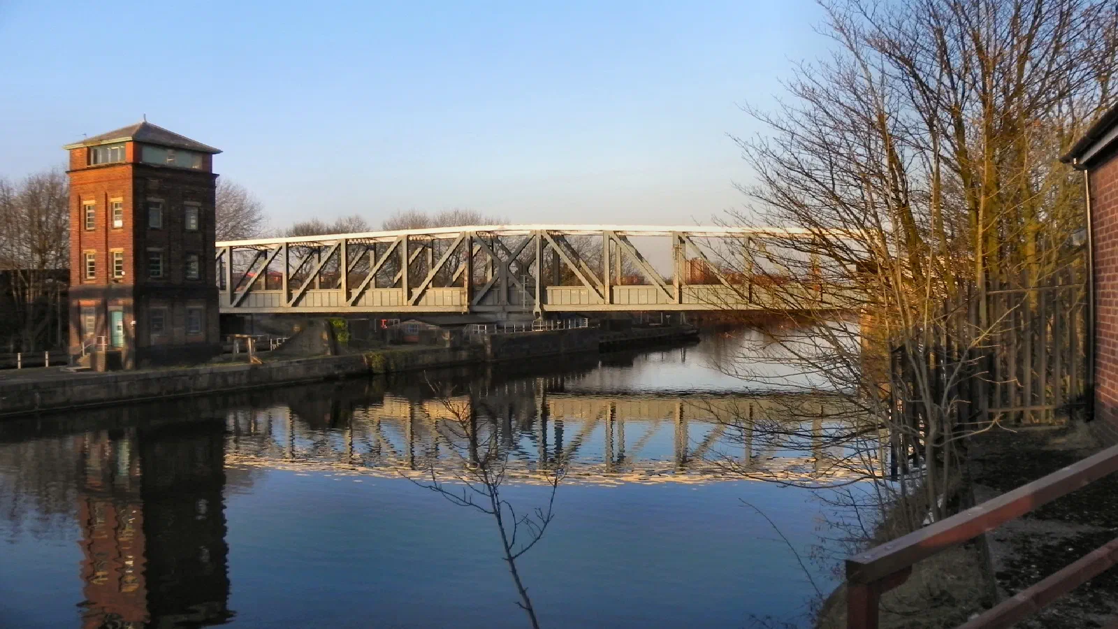 A canal bridge reflecting on calm water beside a brick structure under clear sky. — Barton Swing Bridge & Aqueduct, Manchester