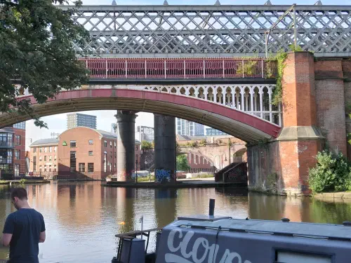 Walkers on a towpath beside a moored narrowboat, under red and grey cast-iron railway viaducts arching across the canal.