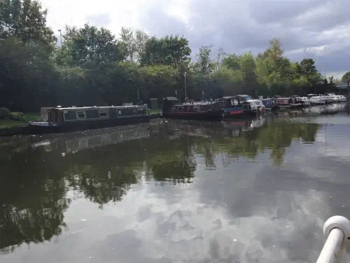 Narrowboats moored along the left bank of a canal, with a road bridge beyond and broken cloud reflected in the water.