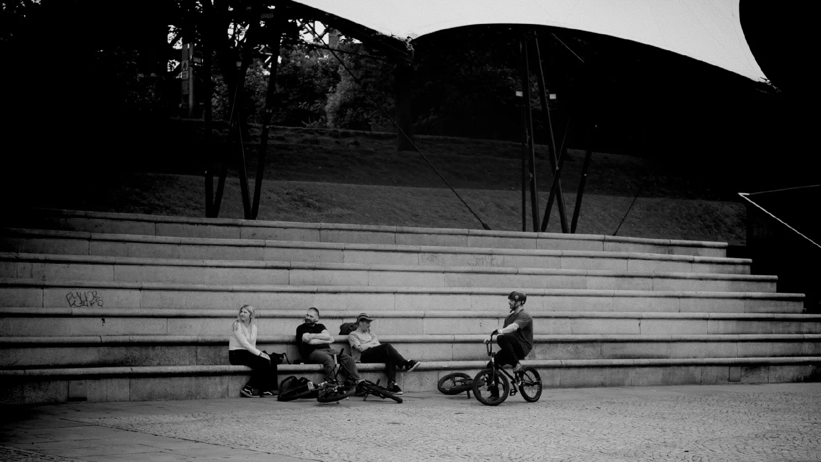 Four people sit on stone steps under a modern canopy while one rides a bicycle in black-and-white. — Castlefield Bowl, Manchester