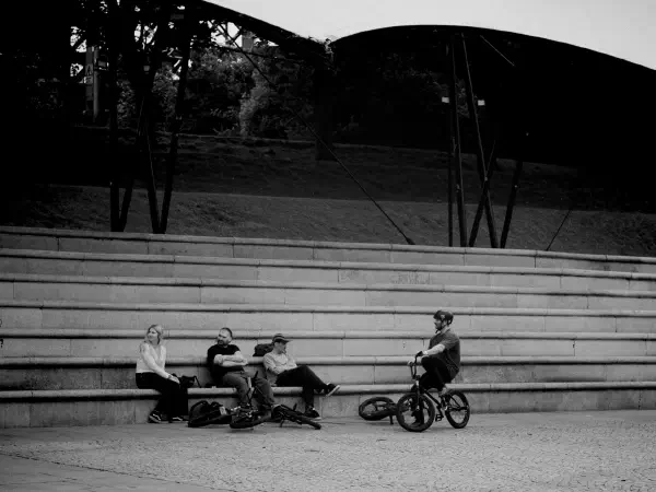 Four people sit on stone steps under a modern canopy while one rides a bicycle in black-and-white. — Castlefield Bowl, Manchester