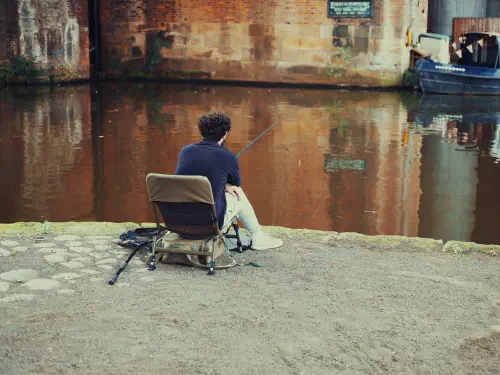 Man fishing by canal wall under overcast skies near moored blue boat.