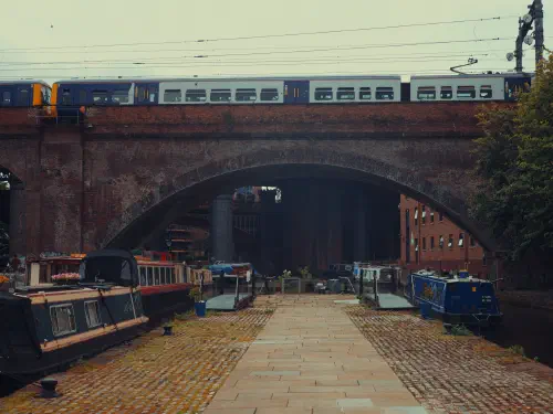 A blue-and-white train crosses a brick bridge over narrowboats docked at a canal path.
