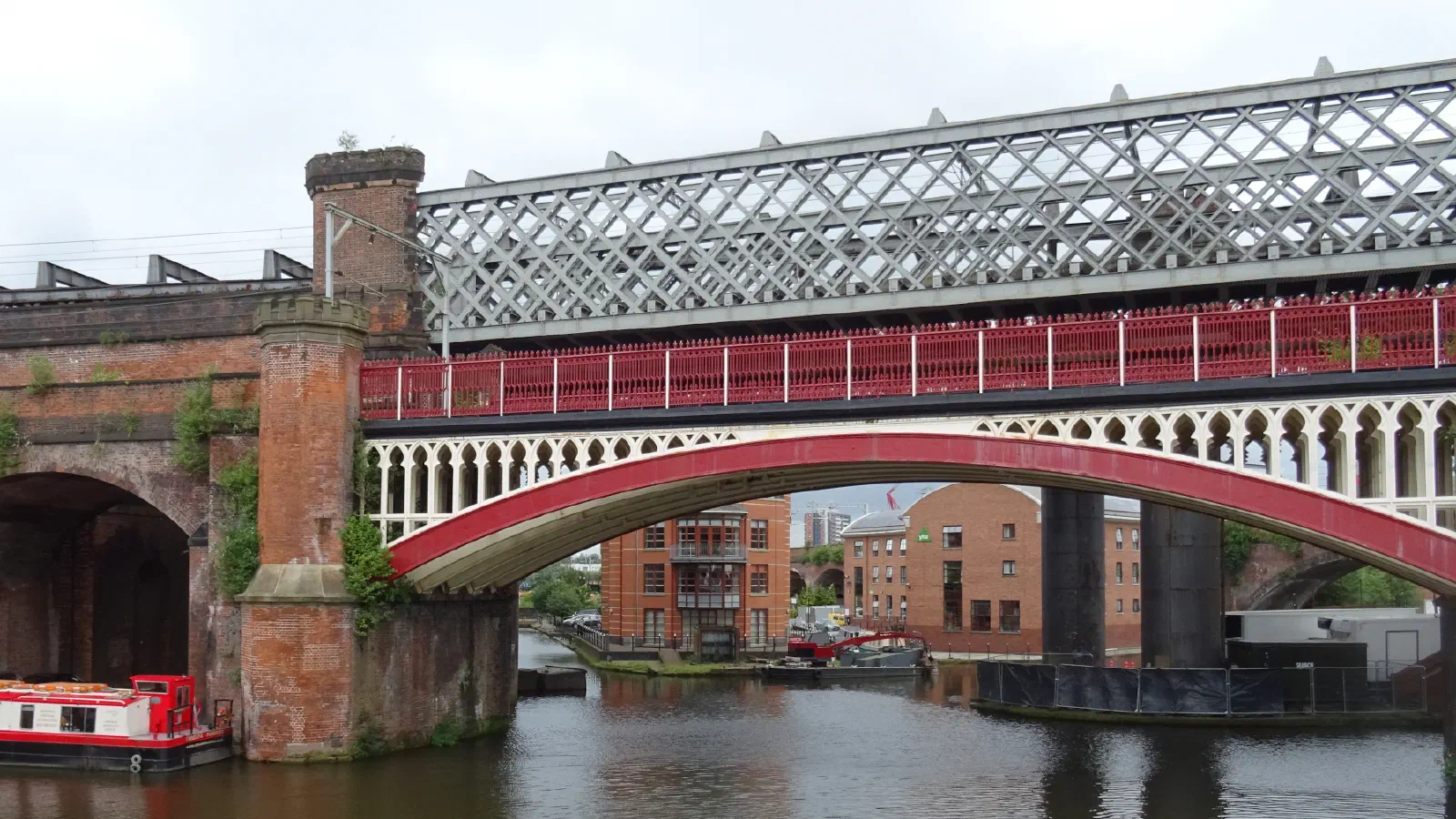 Red railway bridge spans canal; brick arches frame boats and industrial structures under overcast sky. — Castlefield Urban Heritage Park, Manchester