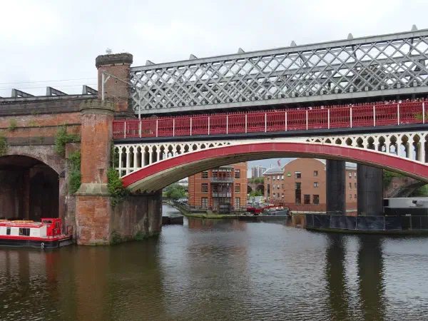 Red railway bridge spans canal; brick arches frame boats and industrial structures under overcast sky. — Castlefield Urban Heritage Park, Manchester