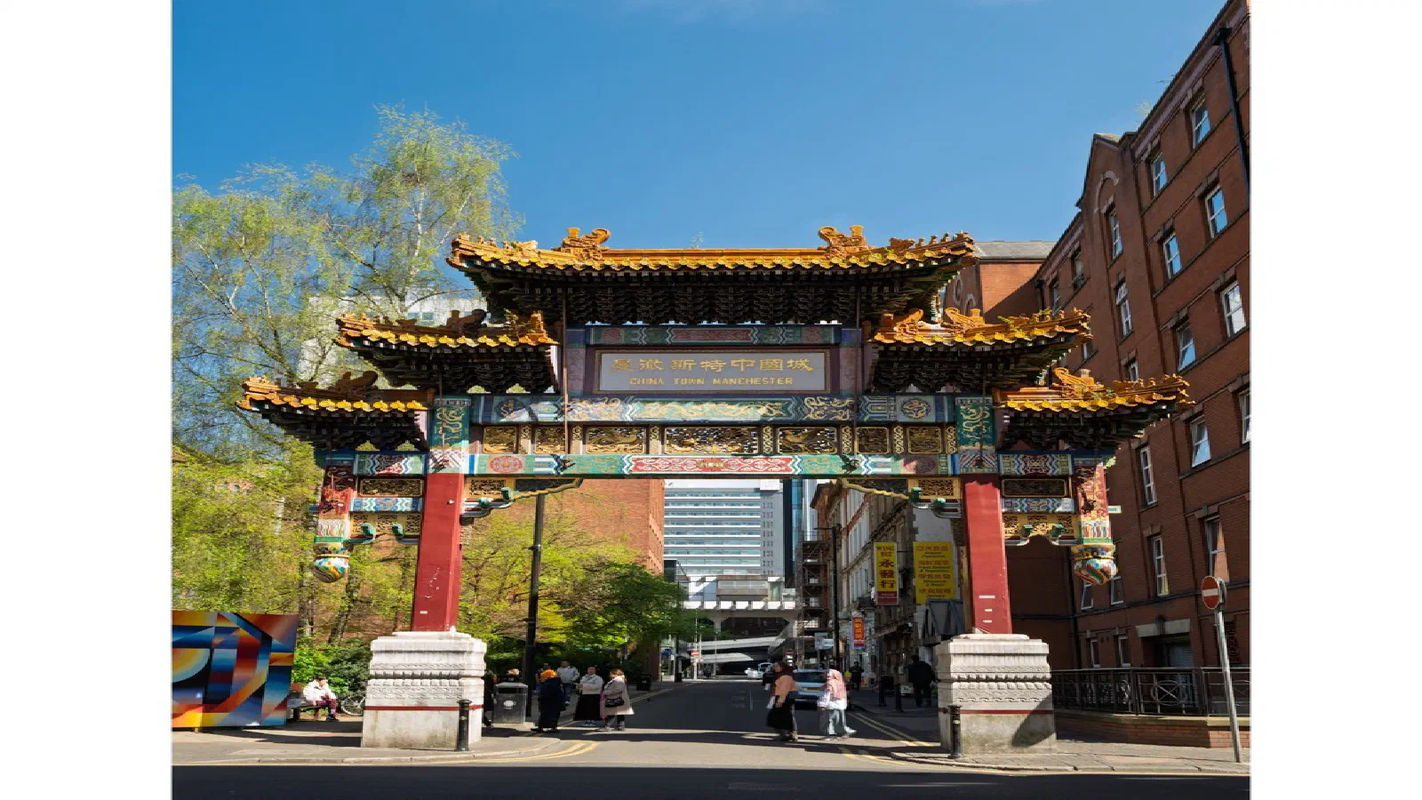 Colorful archway marks Chinatown entrance on sunny day with red-brick building to right. — Chinatown, Manchester
