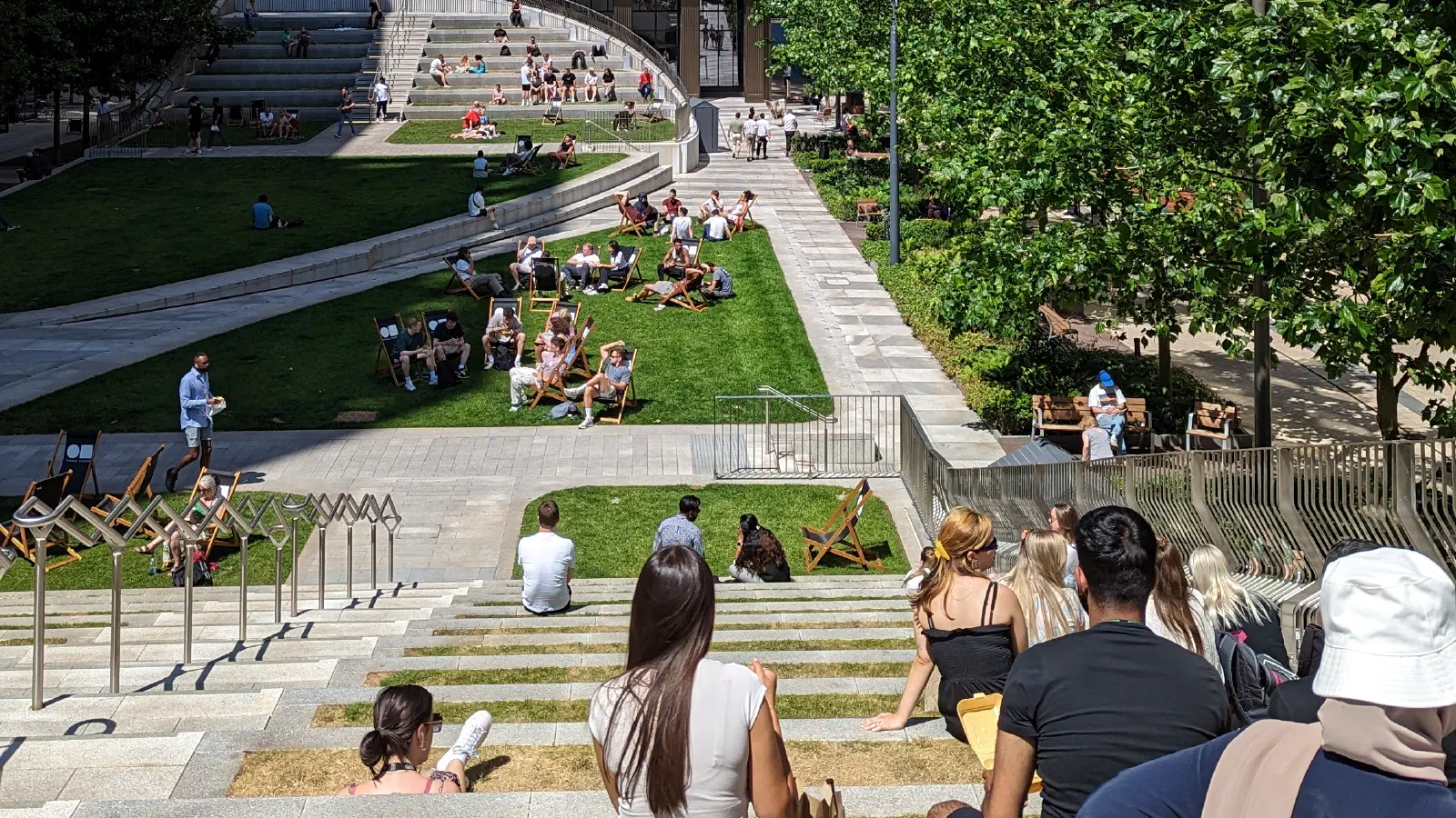 On a sunny day, people relax on steps between modern brick and beige buildings with green grass nearby. — Circle Square (Symphony Park), Manchester