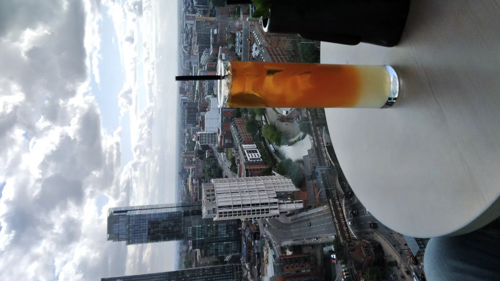 Tall glass orange drink sits on table beside black cup with plant; cityscape below under cloudy sky. — Cloud 23 / 20 Stories, Manchester