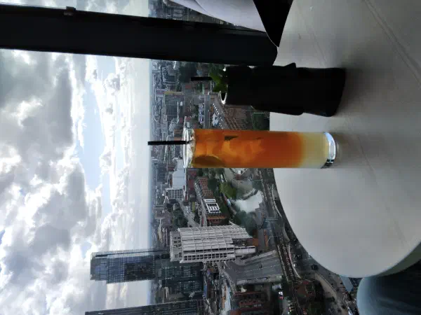 Tall glass orange drink sits on table beside black cup with plant; cityscape below under cloudy sky. — Cloud 23 / 20 Stories, Manchester