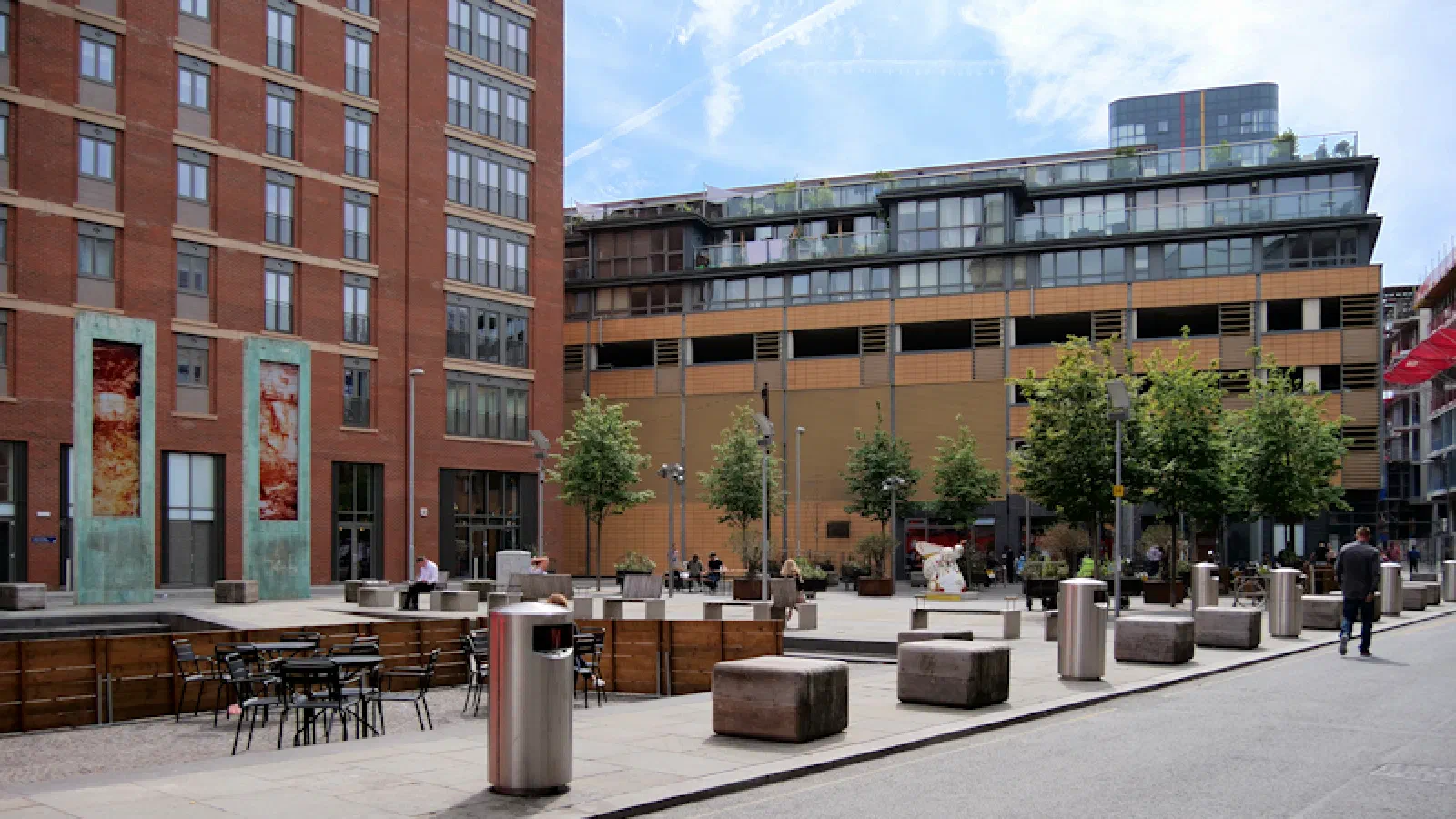 A red brick building stands next to a courtyard with tables, benches, trees, and pedestrians under sunny skies. — Cutting Room Square, Manchester
