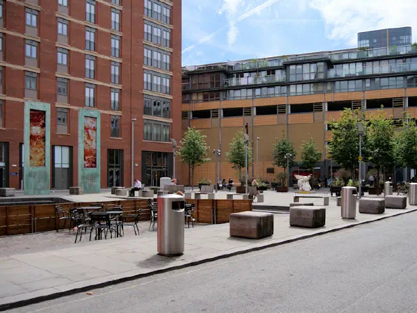 A red brick building stands next to a courtyard with tables, benches, trees, and pedestrians under sunny skies. — Cutting Room Square, Manchester