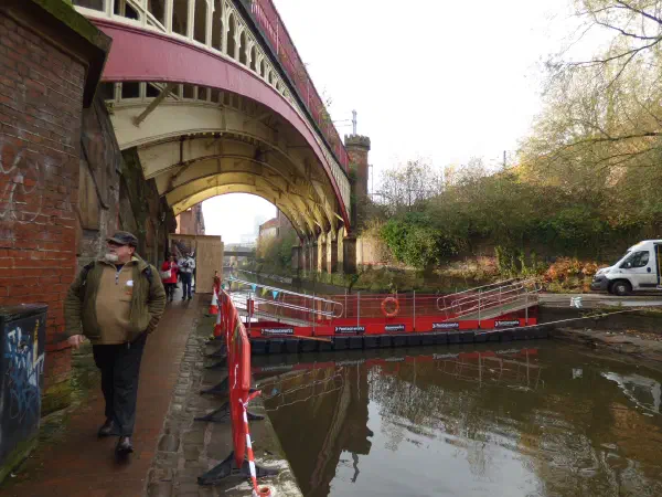 Man walks under red-arched bridge near canal construction site. — Deansgate Locks, Manchester
