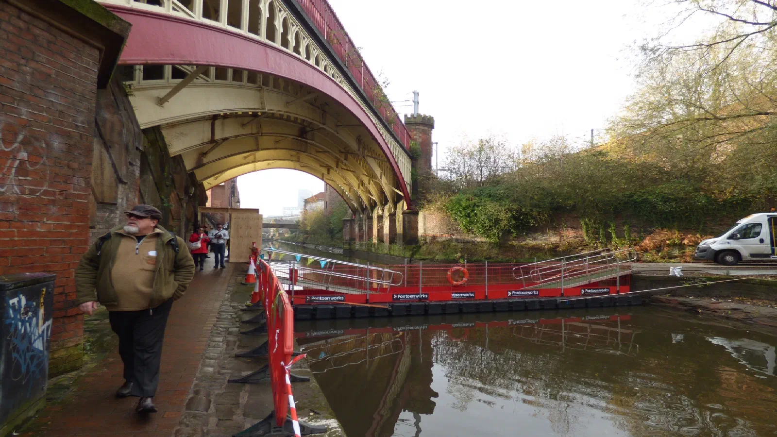 Man walks under red-arched bridge near canal construction site. — Deansgate Locks, Manchester
