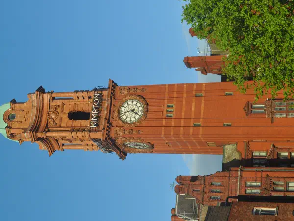Tall brick clock tower named "Kimpton" stands against a clear blue sky with green trees nearby. — The Elizabeth Tower, Manchester