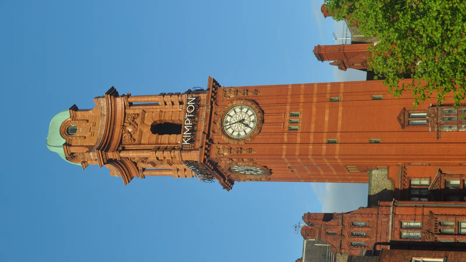 Tall brick clock tower named "Kimpton" stands against a clear blue sky with green trees nearby. — The Elizabeth Tower, Manchester
