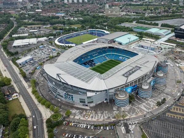 This aerial photo shows an oval-shaped stadium surrounded by parking lots under bright weather. — Etihad Stadium, Manchester