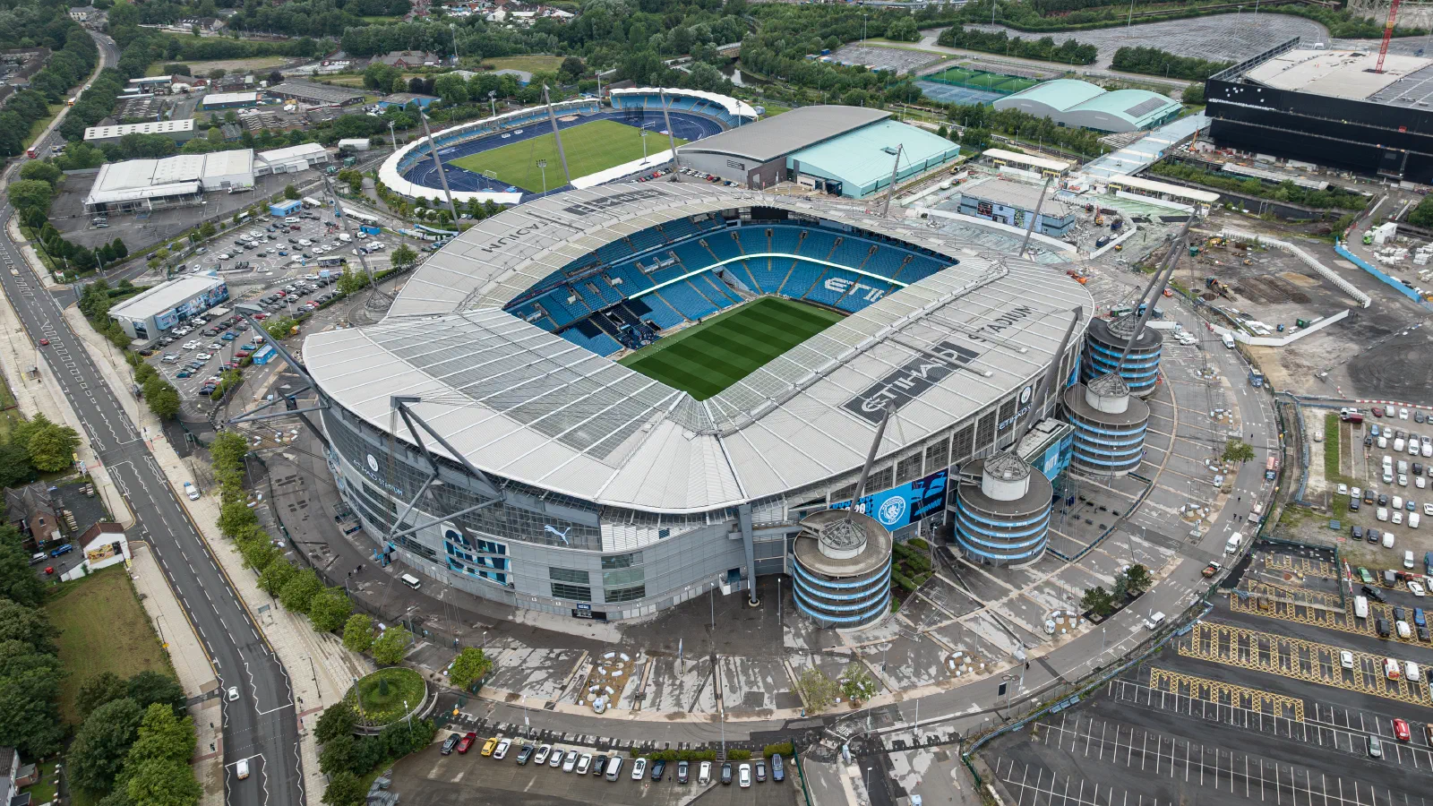 This aerial photo shows an oval-shaped stadium surrounded by parking lots under bright weather. — Etihad Stadium, Manchester