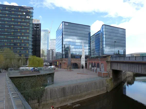 Modern buildings line a riverbank under bright skies, connected by an industrial bridge over calm water. — Greengate Square, Manchester