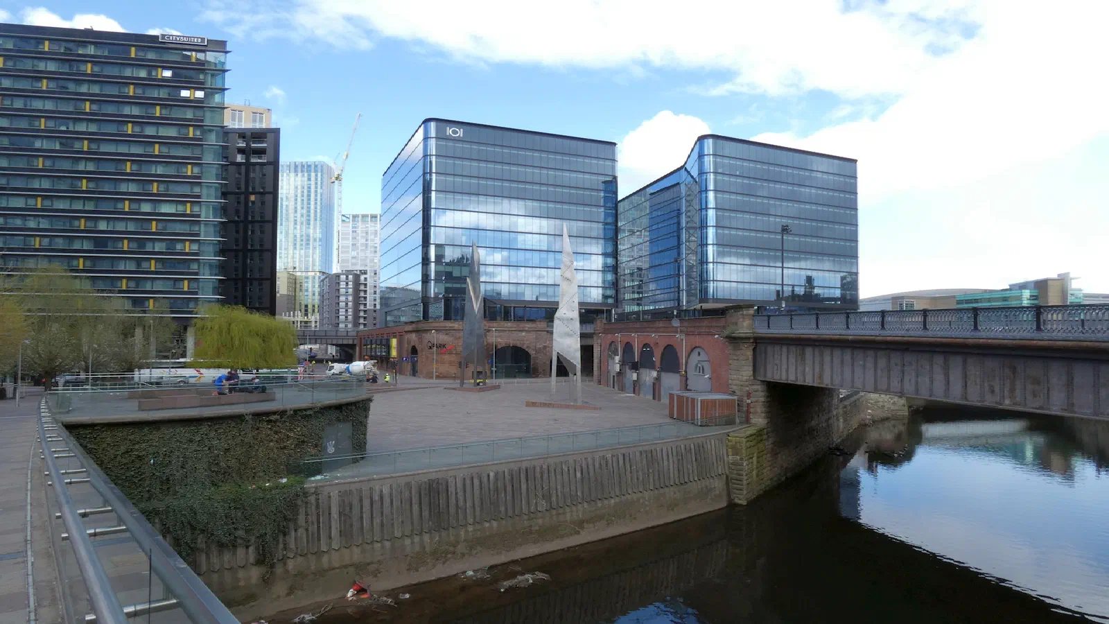 Modern buildings line a riverbank under bright skies, connected by an industrial bridge over calm water. — Greengate Square, Manchester