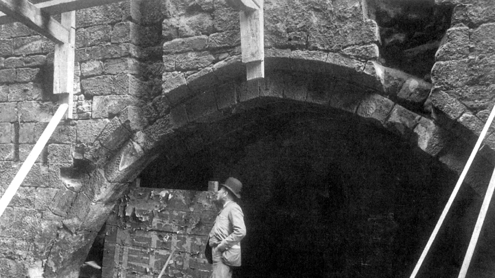 Man stands inside arched stone building under wooden beams, observing a partially ruined brick wall interior. — The Hanging Bridge, Manchester