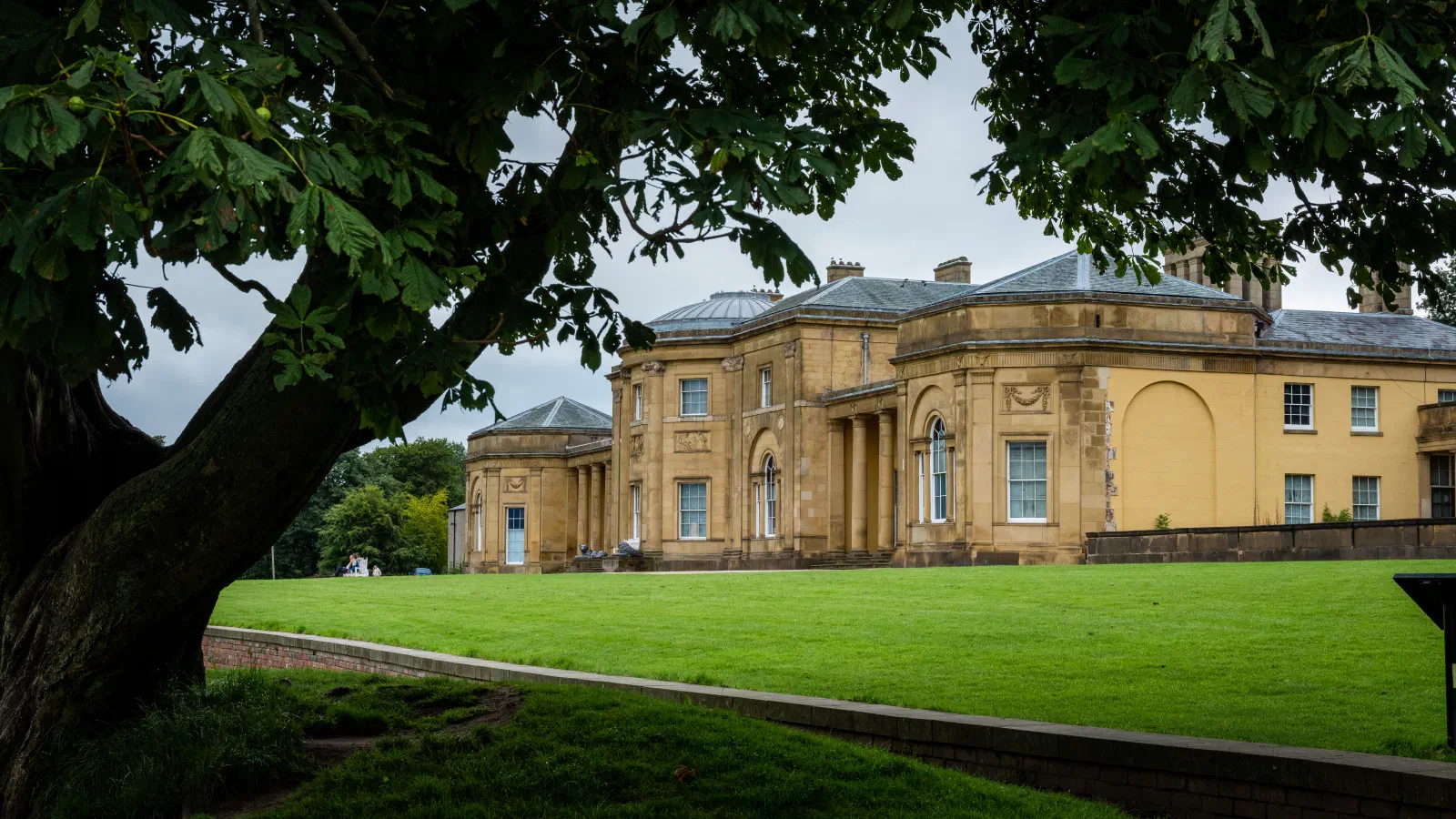 Green lawn stretches before a yellow building under an overcast sky, framed by tree branches. — Heaton Park, Manchester