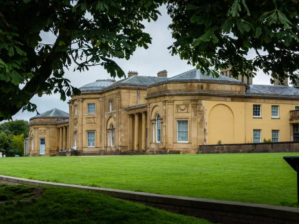 Green lawn stretches before a yellow building under an overcast sky, framed by tree branches. — Heaton Park, Manchester