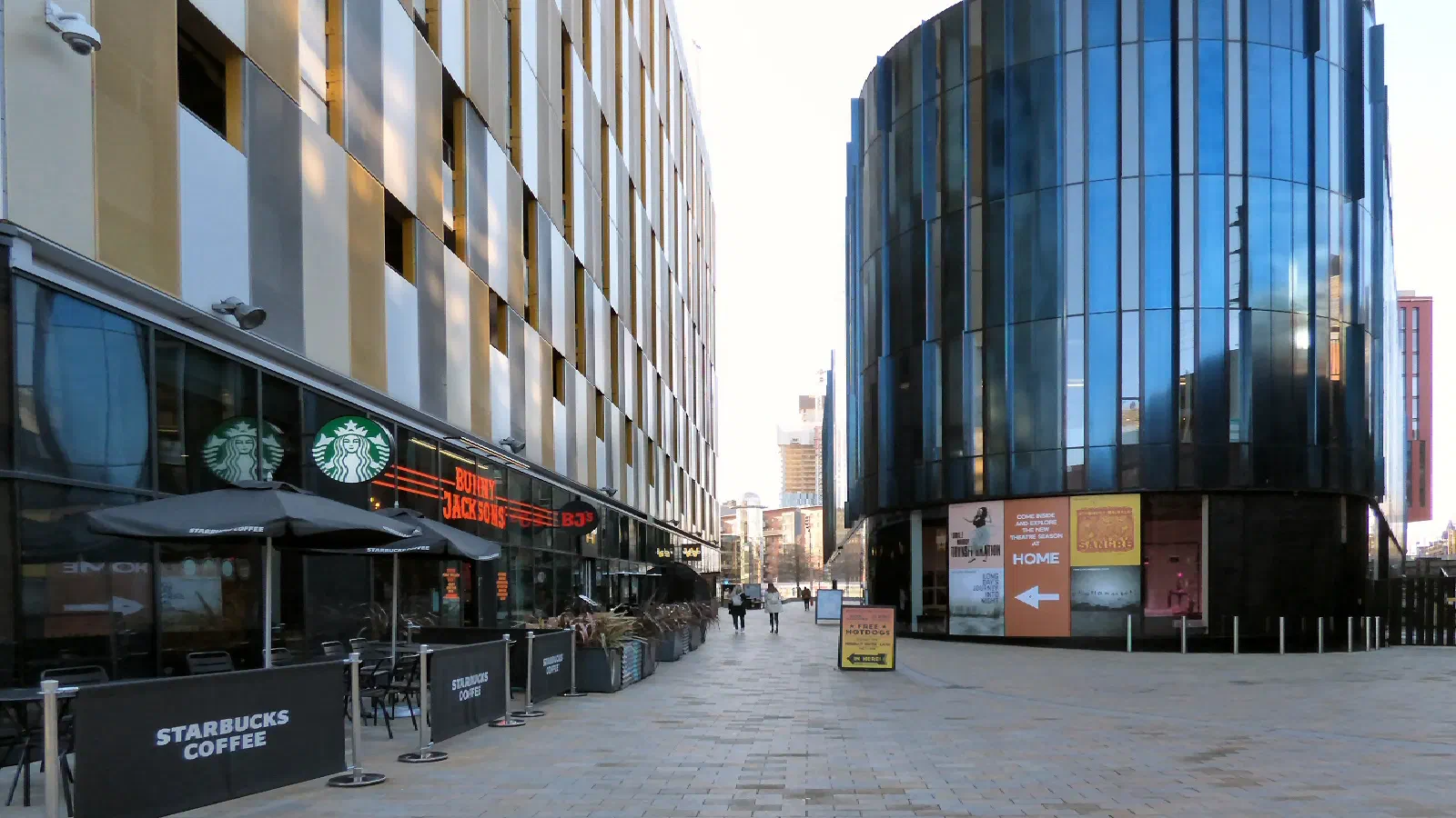 Starbucks outdoor seating lines a paved plaza beside reflective glass building under clear sky. — HOME Manchester, Manchester