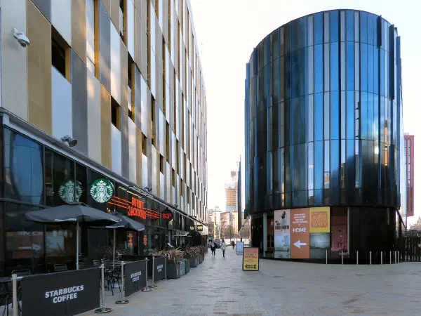 Starbucks outdoor seating lines a paved plaza beside reflective glass building under clear sky. — HOME Manchester, Manchester