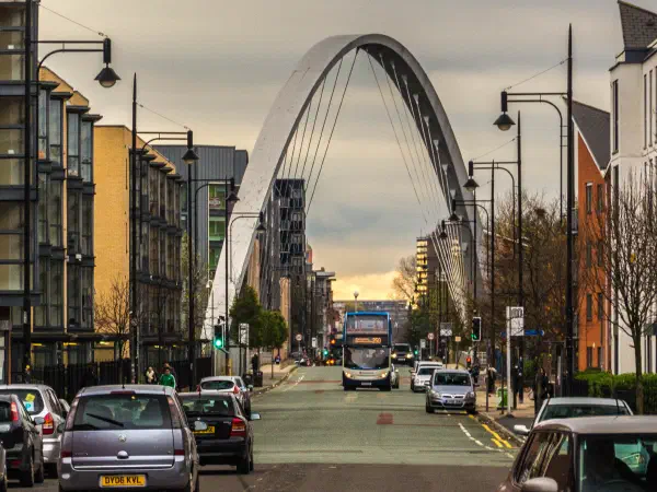 Modern arch bridge spans urban street lined with cars under cloudy skies. — Hulme Arch Bridge, Manchester