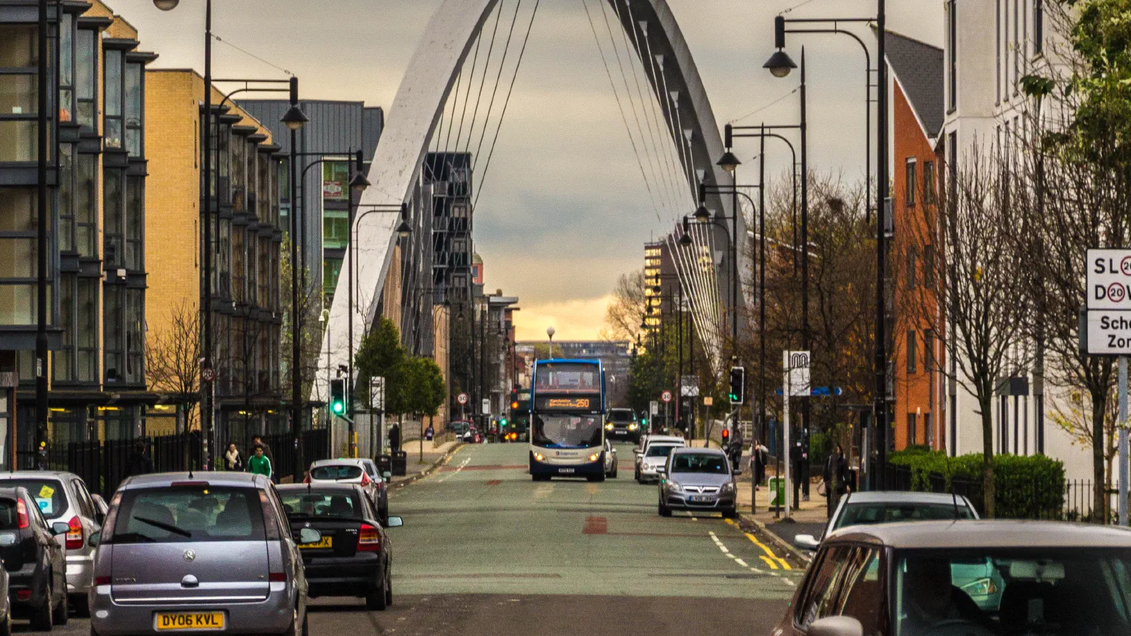 Modern arch bridge spans urban street lined with cars under cloudy skies. — Hulme Arch Bridge, Manchester