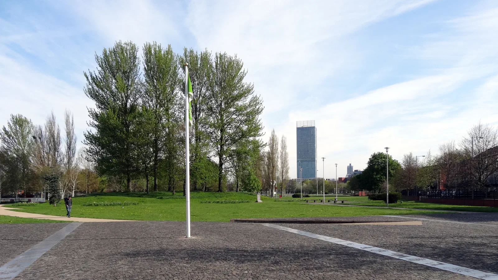 Green flag on cobblestone plaza near trees, tall building, blue sky. — Hulme Park, Manchester