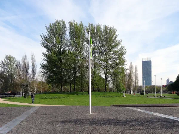 Green flag on cobblestone plaza near trees, tall building, blue sky. — Hulme Park, Manchester