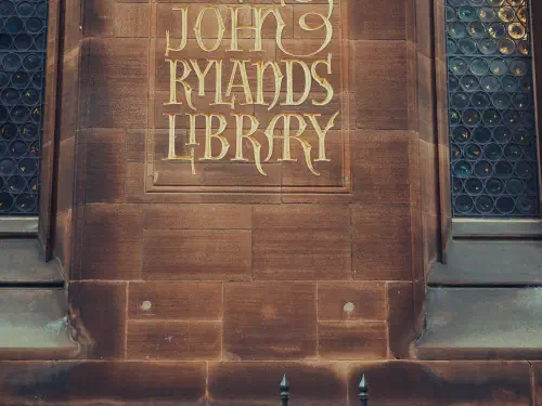 Golden lettering on stone wall reads 'The John Rylands Library,' flanked by stained glass windows with gothic arches.
