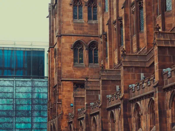 People walk on a stone path beside tall red-brick Gothic buildings near a modern blue glass structure under overcast skies. — John Rylands Library, Manchester