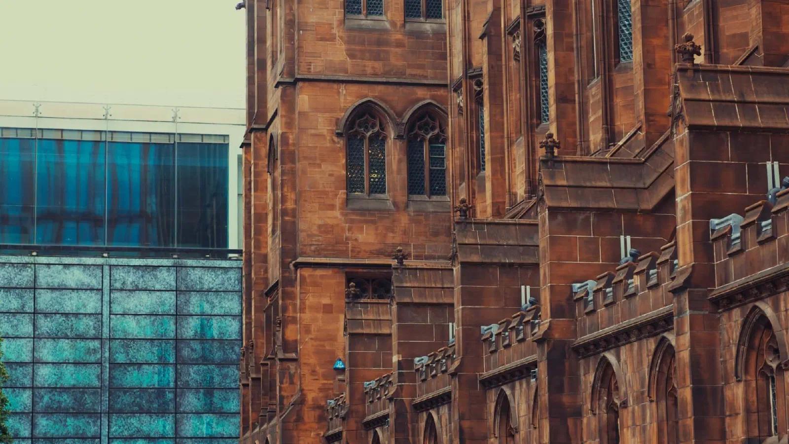 People walk on a stone path beside tall red-brick Gothic buildings near a modern blue glass structure under overcast skies. — John Rylands Library, Manchester