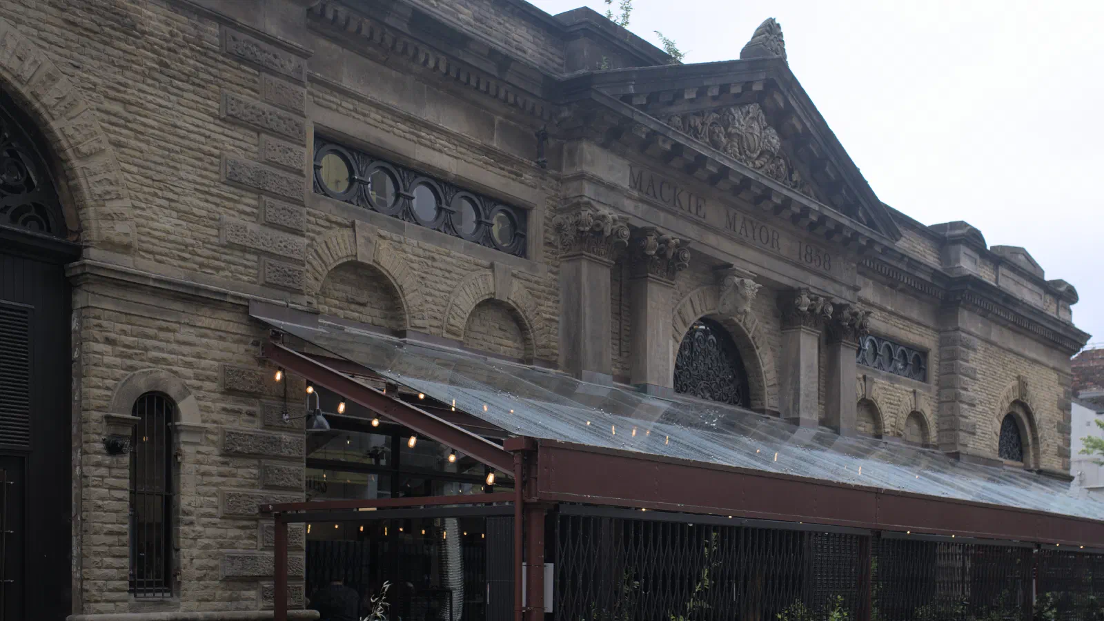 The historic stone building with a glass-roofed terrace, featuring inscribed text above arched windows under an overcast sky. — Mackie Mayor, Manchester
