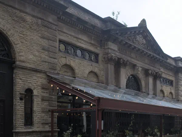 The historic stone building with a glass-roofed terrace, featuring inscribed text above arched windows under an overcast sky. — Mackie Mayor, Manchester