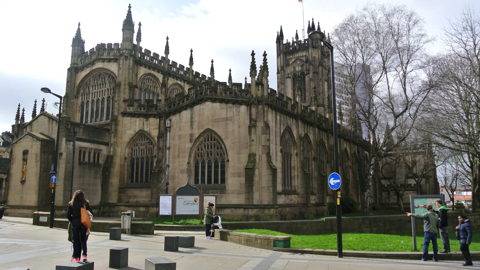 People walk around stone cathedral surrounded by bare trees on cloudy day. — Manchester Cathedral, Manchester