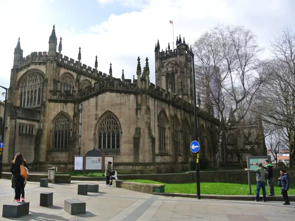 People walk around stone cathedral surrounded by bare trees on cloudy day. — Manchester Cathedral, Manchester