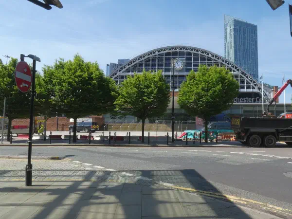 Arched glass and steel roof behind trees, with construction vehicles on the road under a clear blue sky. — Manchester Central (Convention Centre), Manchester