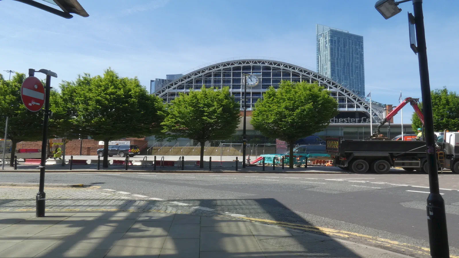 Arched glass and steel roof behind trees, with construction vehicles on the road under a clear blue sky. — Manchester Central (Convention Centre), Manchester