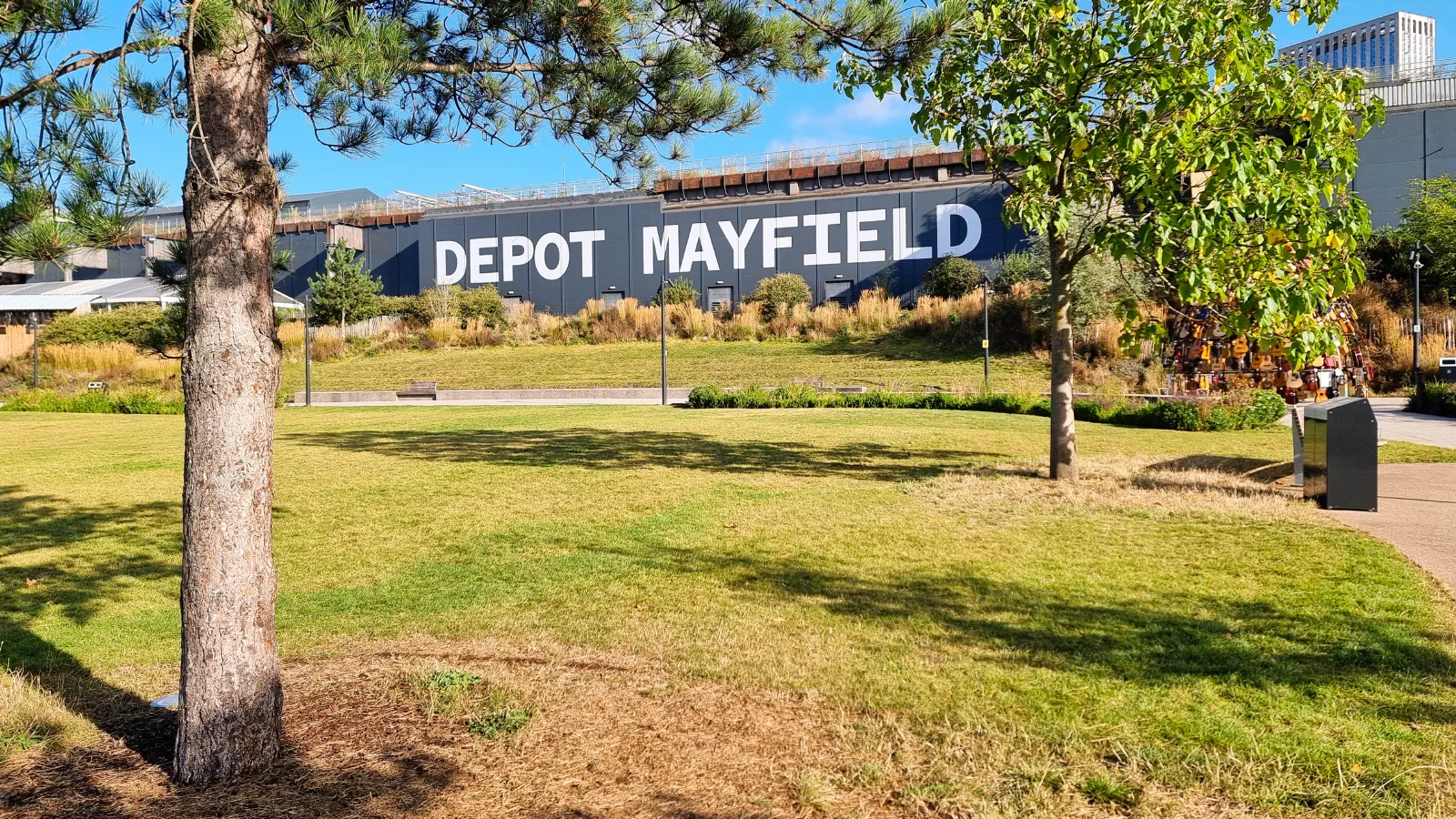 Sunlit grassy field bordered by trees; "DEPOT MAYFIELD" on a building behind under blue sky — Mayfield Depot, Manchester
