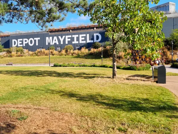 Sunlit grassy field bordered by trees; "DEPOT MAYFIELD" on a building behind under blue sky — Mayfield Depot, Manchester