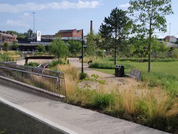 Sunny park scene with grassy field, walking paths, benches, trees, and people relaxing near industrial buildings in background. — Mayfield Park, Manchester