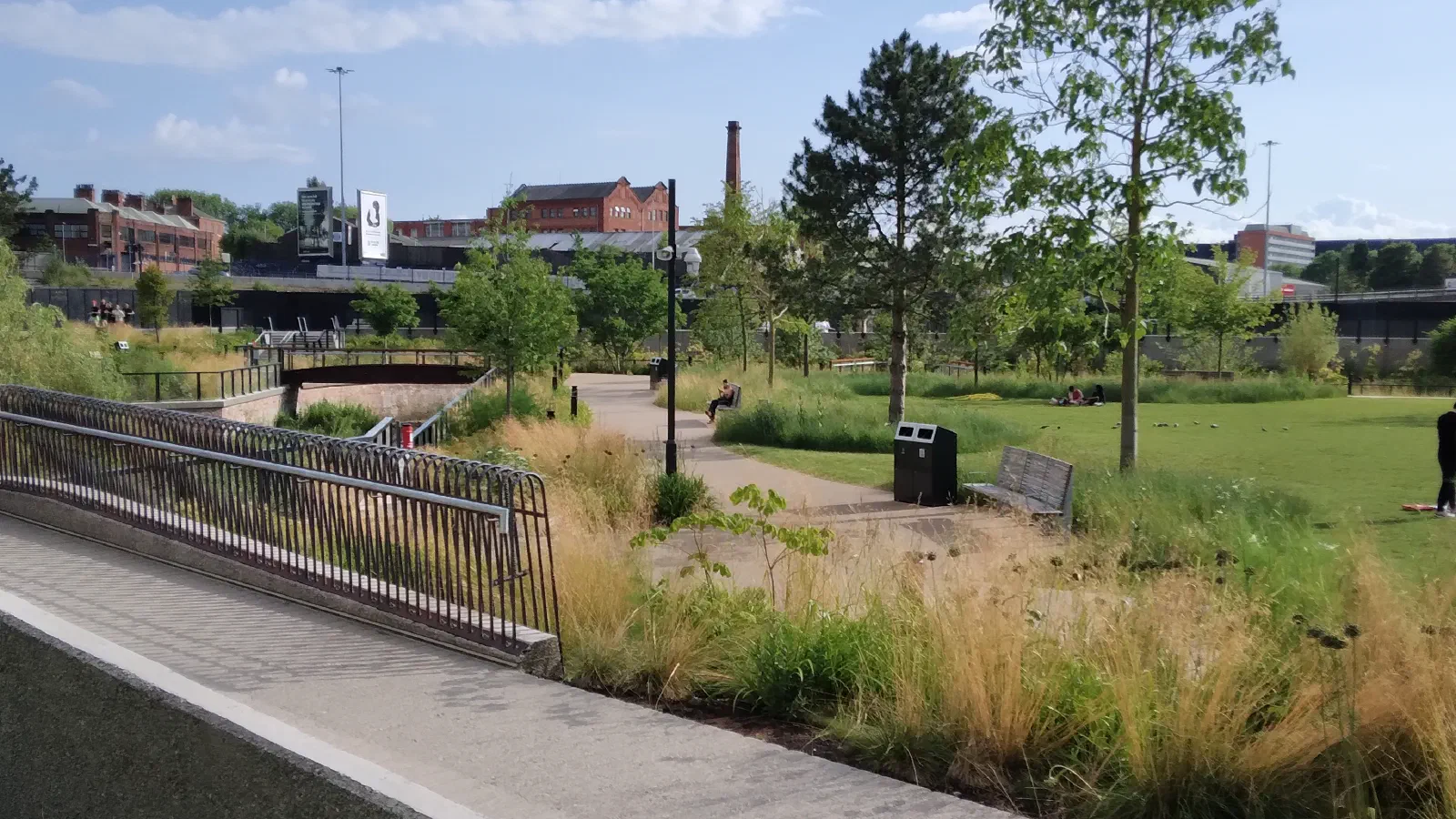 Sunny park scene with grassy field, walking paths, benches, trees, and people relaxing near industrial buildings in background. — Mayfield Park, Manchester