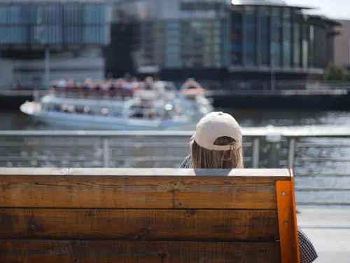 A person sits on a wooden bench overlooking a river with a blurred boat and modern building under bright daylight.